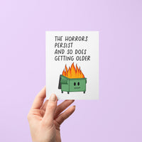 Funny birthday card with illustration of a flaming dumpster and the words “The horrors persist and so does getting older” in bold text, blank inside with kraft envelope.