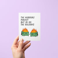 Humorous holiday card with two flaming dumpster characters holding hands, decorated with string lights, and the text “The horrors persist but so do the holidays.”