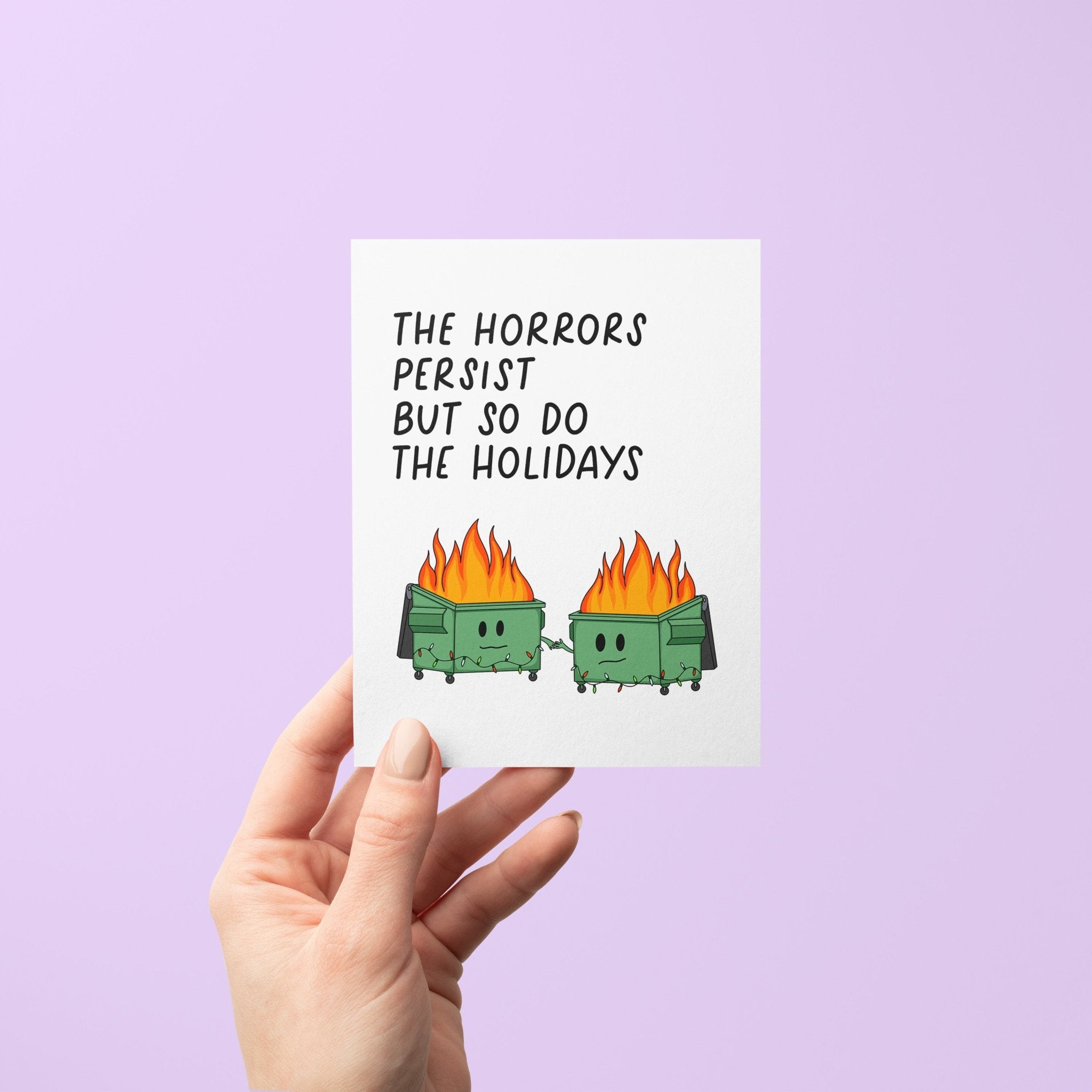 Humorous holiday card with two flaming dumpster characters holding hands, decorated with string lights, and the text “The horrors persist but so do the holidays.”