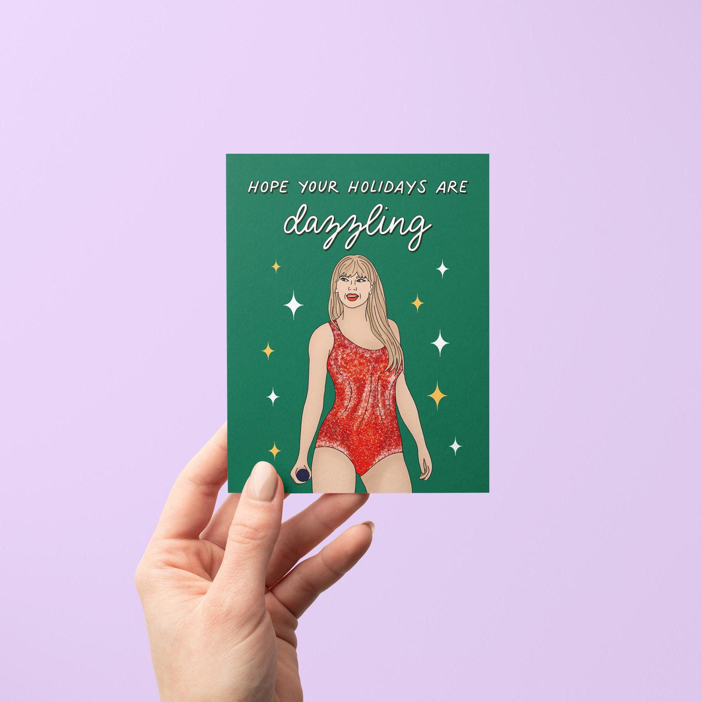 This card features an illustration of Tay in a red version of her Lover era Eras Tour bodysuit surrounded by stars against a green background. The greeting in mixed script and sans serif hand lettering reads &quot;hope your holidays are dazzling.&quot;