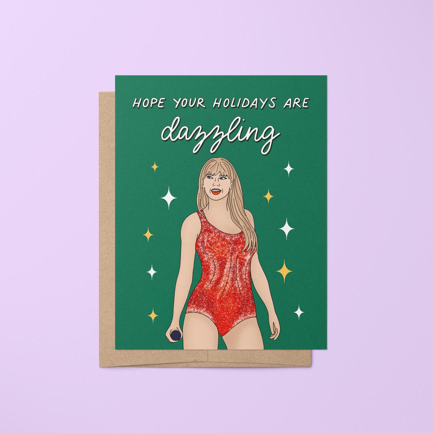 This card features an illustration of Tay in a red version of her Lover era Eras Tour bodysuit surrounded by stars against a green background. The greeting in mixed script and sans serif hand lettering reads &quot;hope your holidays are dazzling.&quot;