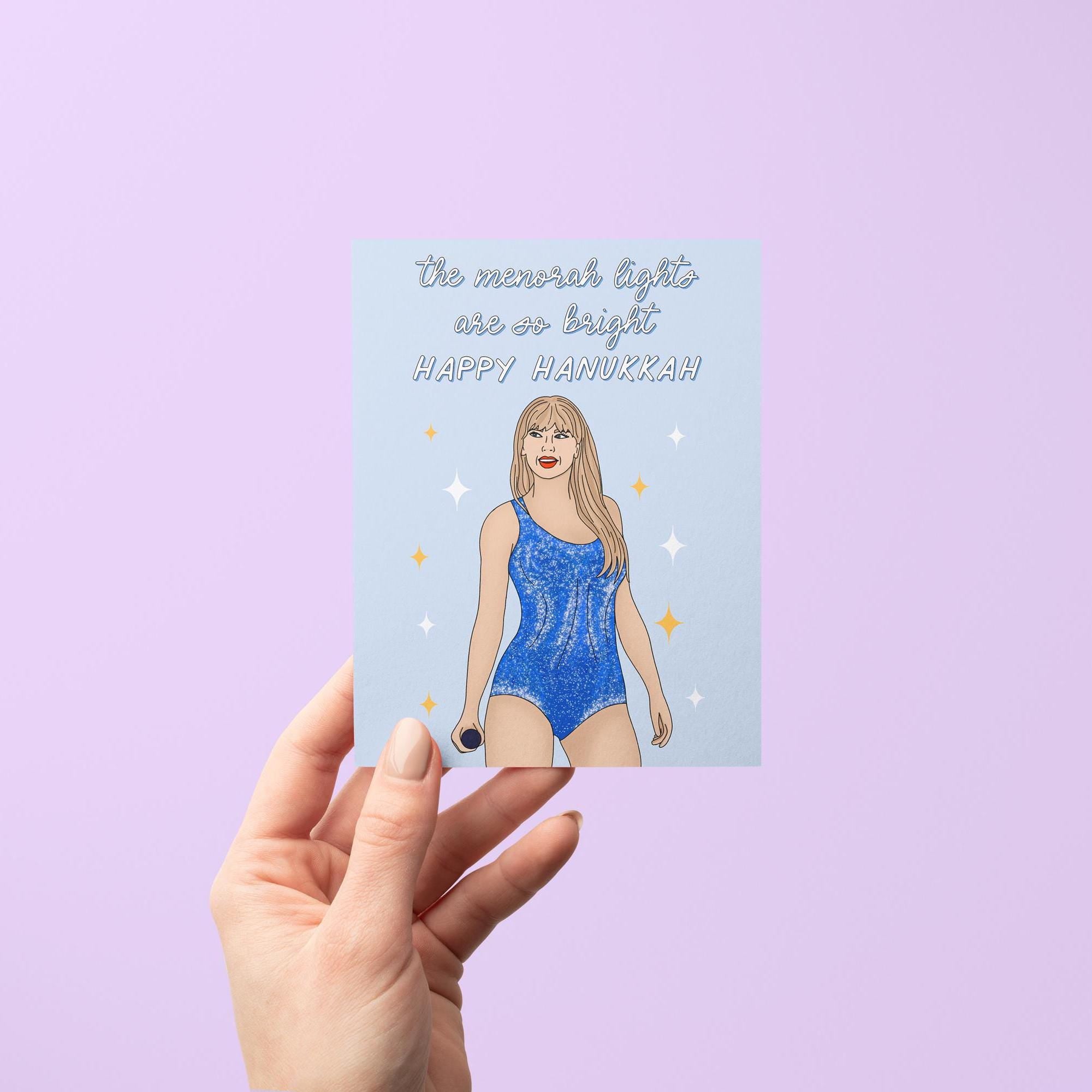 This Swiftie Hanukkah card features an illustration of Tay in a blue version of her Lover era Eras Tour bodysuit surrounded by stars against a light blue background. The greeting reads &quot;the menorah lights are so bright. Happy Hanukkah.&quot;