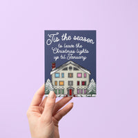 This Swiftie holiday card features an illustration of the Lover House decked out in Christmas lights and dusted with snow. The greeting reads &quot;&#39;Tis the season to leave the Christmas lights up till January.&quot;