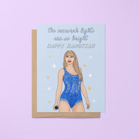 This Swiftie Hanukkah card features an illustration of Tay in a blue version of her Lover era Eras Tour bodysuit surrounded by stars against a light blue background. The greeting reads &quot;the menorah lights are so bright. Happy Hanukkah.&quot;