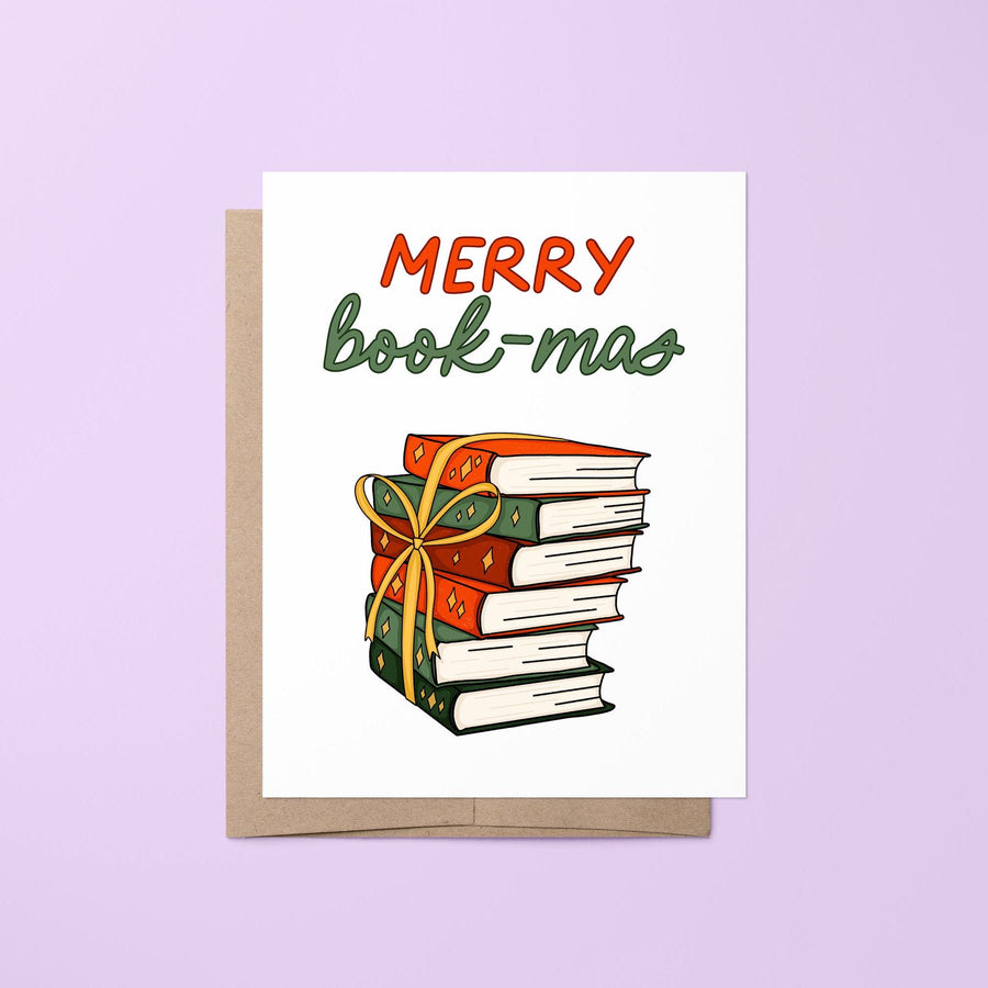 This card features the hand lettered greeting &quot;Merry Book-mas&quot; in red and green shades and features a hand drawn illustration of a stack of books in festive green and red shades with golden stars tied together with a gold ribbon bow.