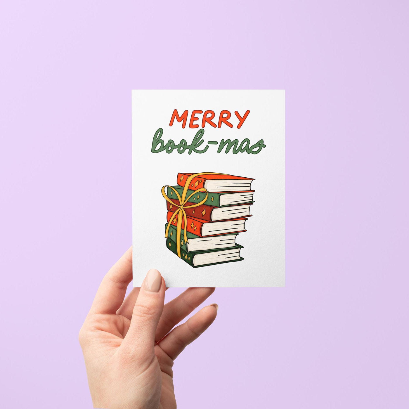 This card features the hand lettered greeting &quot;Merry Book-mas&quot; in red and green shades and features a hand drawn illustration of a stack of books in festive green and red shades with golden stars tied together with a gold ribbon bow.