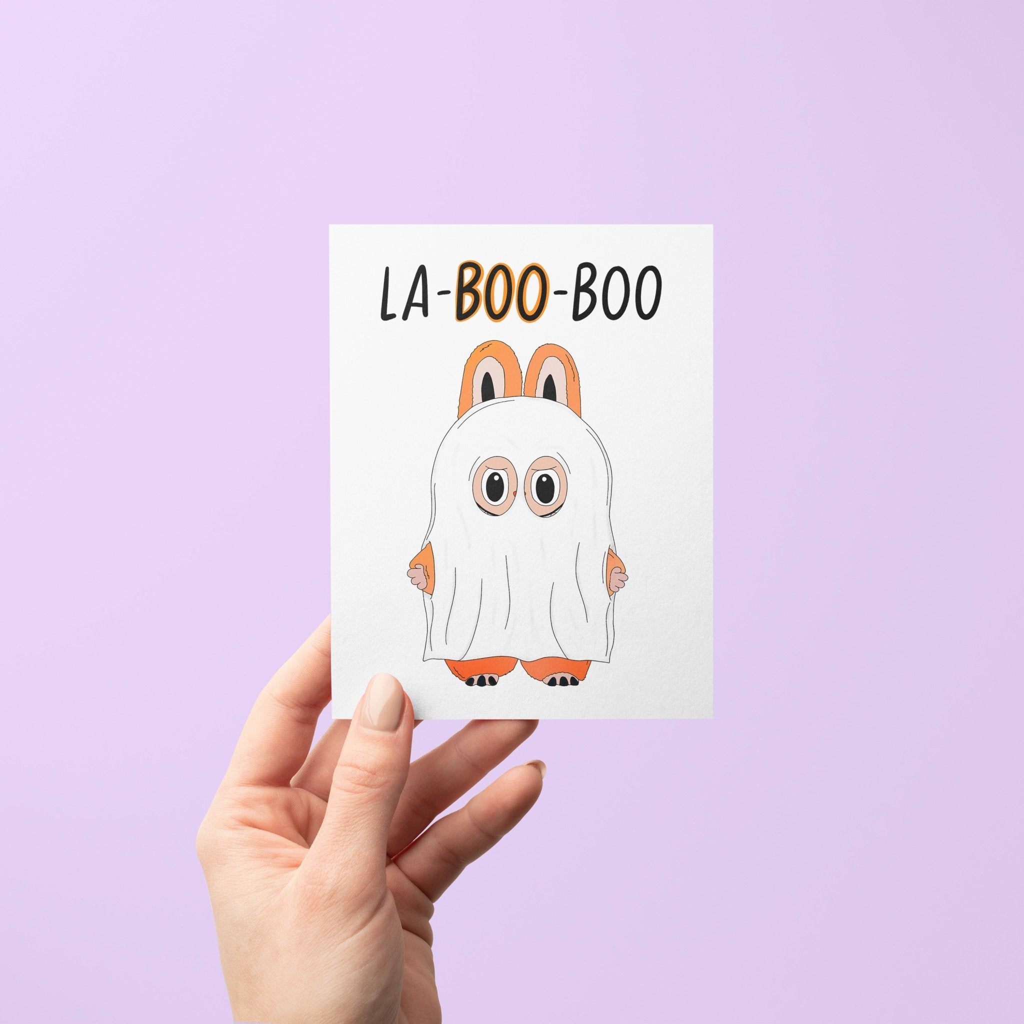 Halloween greeting card with the words “La-BOO-boo” and an illustration of a cute monster in a ghost costume with bunny ears.