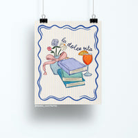 Illustrated art print featuring pastel books, a vase of flowers with a ribbon, and an Aperol spritz with the handwritten text "la dolce vita" on a striped background.